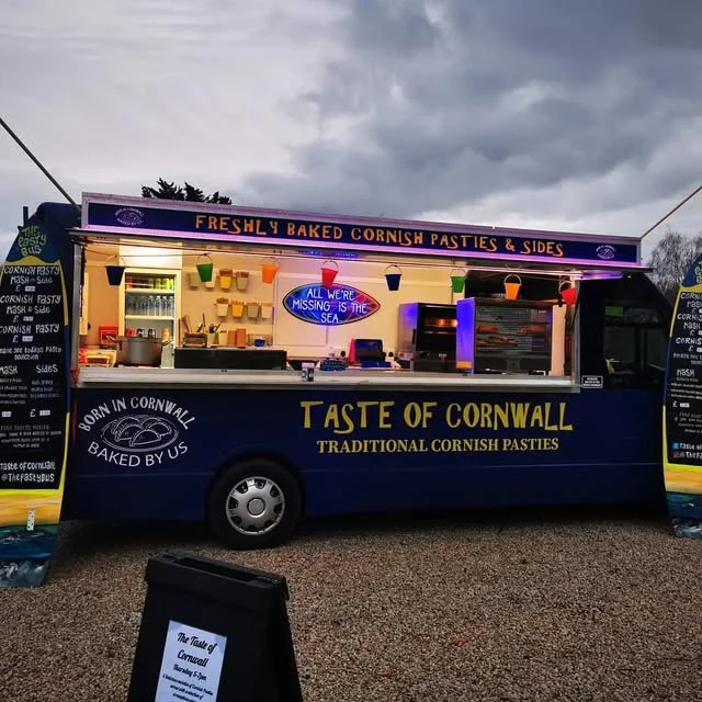 The bus set up at dusk, yellow Taste of Cornwall lettering along the navy side and the hatch lit from within.